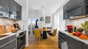 Uma cozinha moderna com eletrodomésticos de aço inoxidável, um espaço de refeições com uma mesa redonda e cadeiras amarelas, e vista da cidade através de grandes janelas.