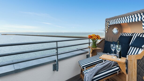 Varanda com vista para o mar, mesa de madeira com dois copos e uma garrafa, e almofadas listradas.