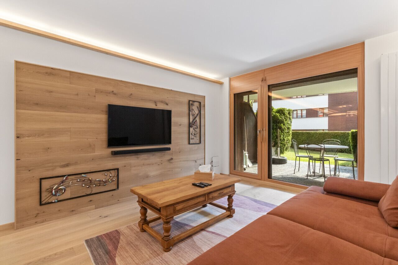 A modern living room with a wooden wall, a flat-screen TV, a wooden coffee table, and a large window with a view of a garden and building.