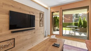 A modern living room with a flat-screen TV mounted on a wooden wall, a wooden coffee table, and a large sliding glass door leading to an outdoor patio with a table and chairs.