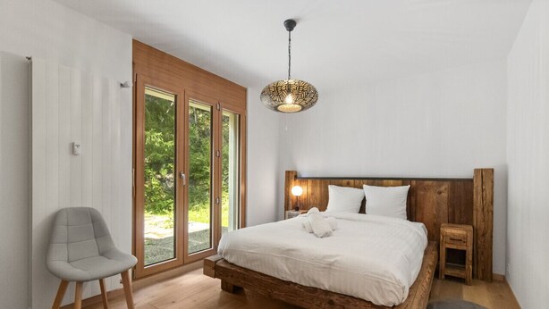 A bedroom with a wooden bed, a grey chair, a small bedside table, and a large window with a view of greenery.
