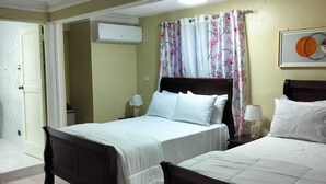 Une chambre d’hôtel avec deux lits, un ventilateur de plafond, un appareil de climatisation, une fenêtre avec des rideaux à motifs floraux et un tableau encadré au mur.