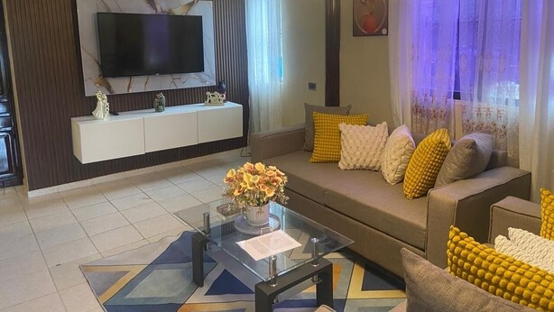 Un salon moderne avec un téléviseur à écran plat, un canapé orné de coussins jaunes et blancs, une table basse en verre sur laquelle est posé un vase de fleurs, et un tapis à motifs.