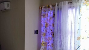 Une pièce aux murs de couleur beige, avec un climatiseur blanc, un rideau de douche orné de motifs floraux et une fenêtre agrémentée d’un rideau à fleurs.