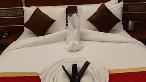 Cama bem arrumada com colcha vermelha e branca, duas almofadas marrons e uma toalha branca dobrada sobre a cama.