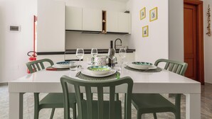 A dining area with a white table set for four, green chairs, and a kitchen in the background. - 4065 Kalès Holiday by Barbarhouse (Otranto)