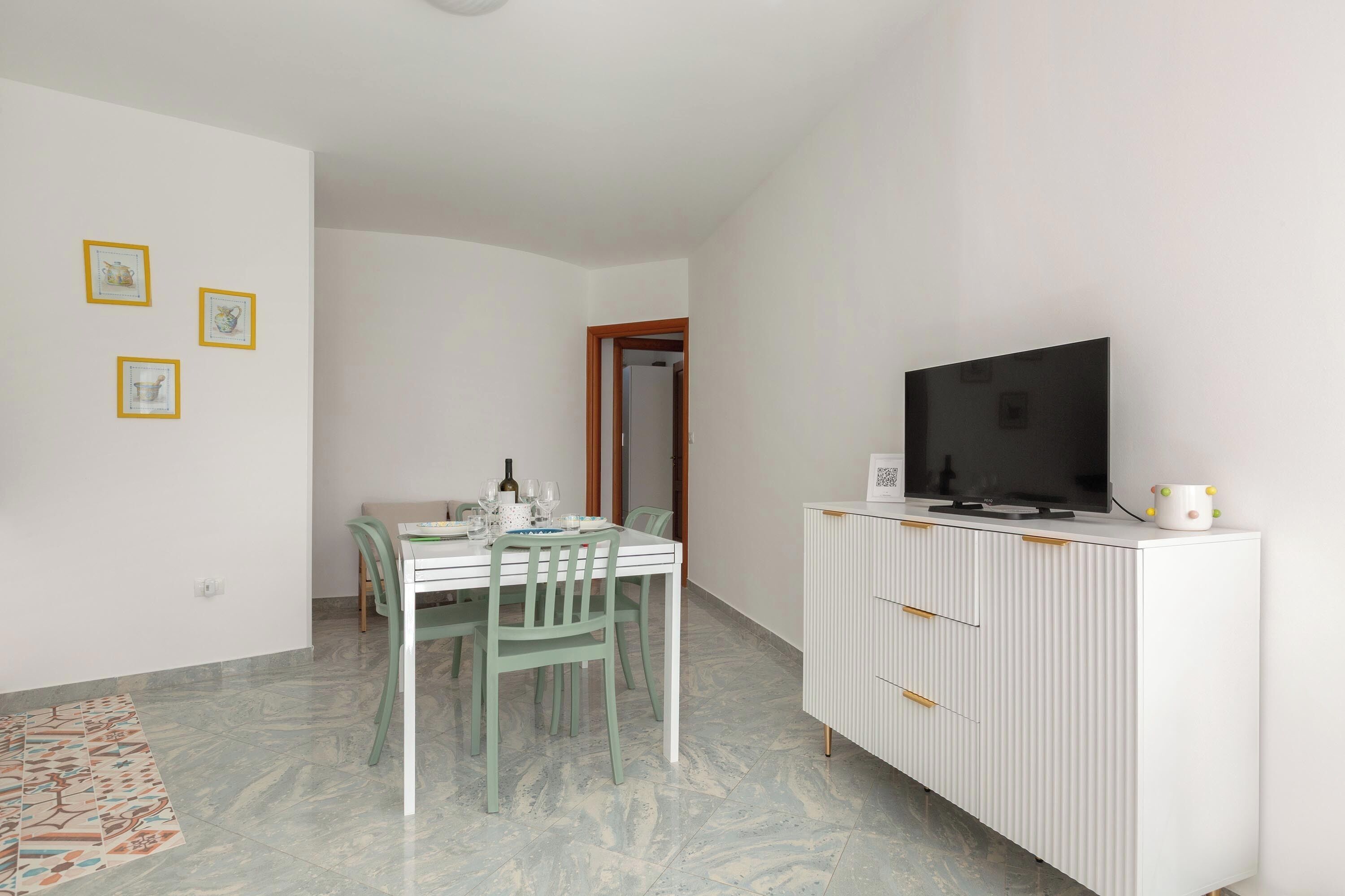 Un área de comedor con una mesa blanca y sillas verdes, un armario blanco con un televisor, y tres cuadros en la pared.