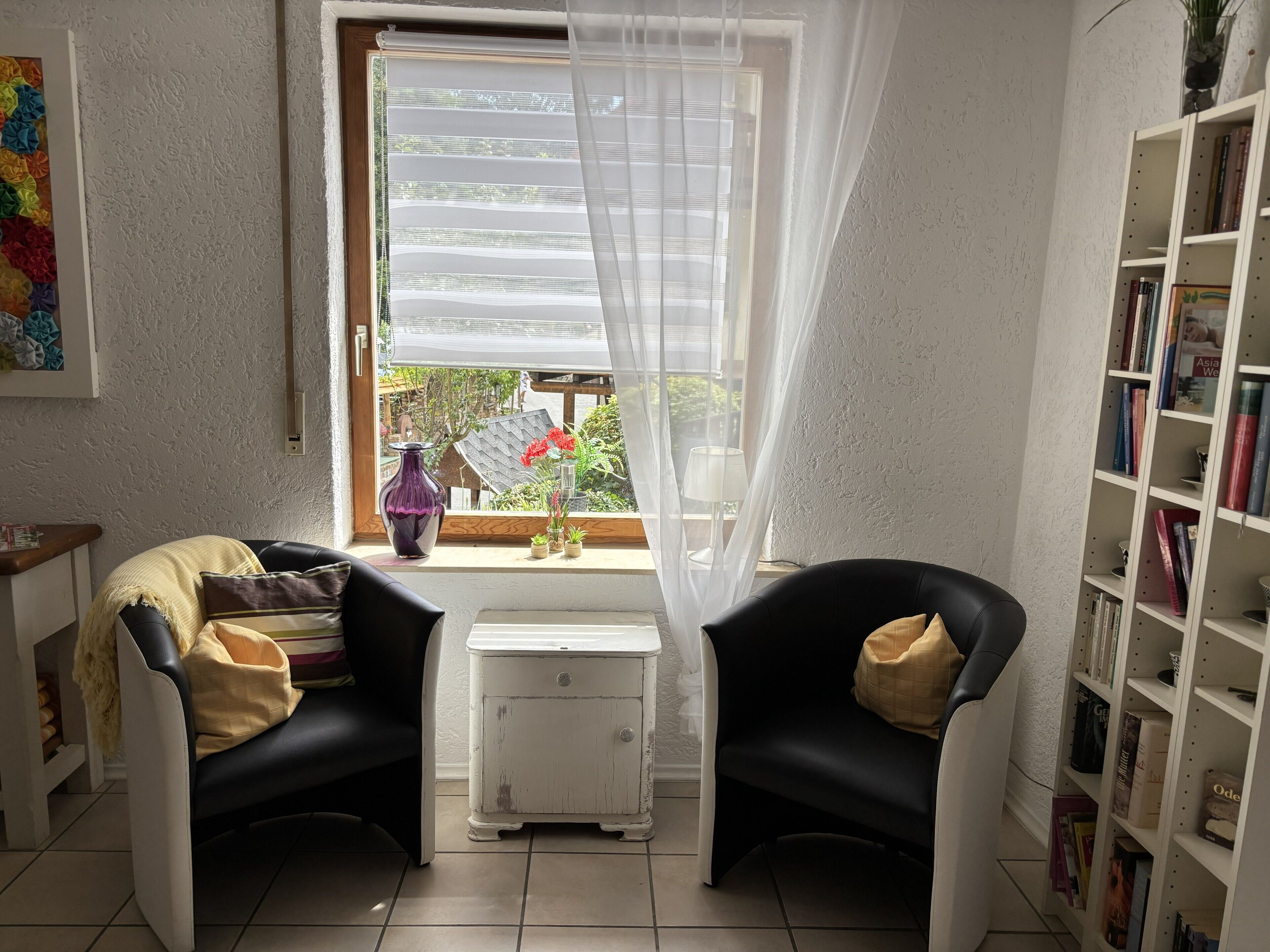 Een kamer met twee zwarte stoelen, een witte bijzettafel, een boekenplank en een raam met dunne gordijnen.