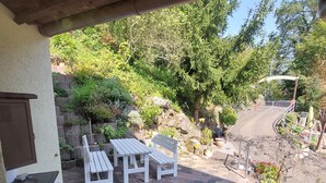 An outdoor patio with a brick floor, white furniture, and a view of a garden and street. - Ferienhaus "gängelbach" Auszeit im Grünen bei Bachrauschen! (Weinheim)