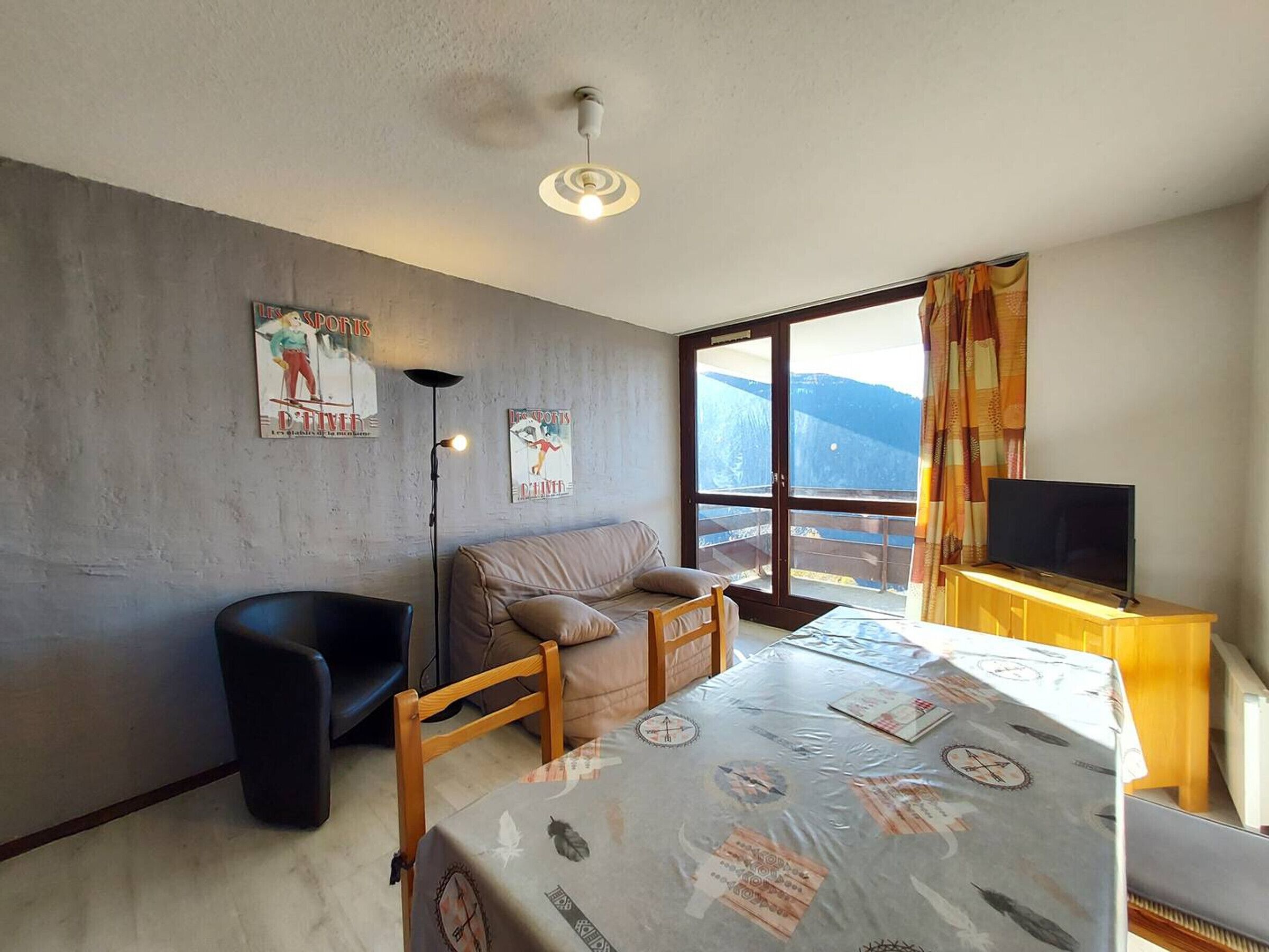 A modern room with a sofa, a small table, a TV on a wooden stand, and a large window with a view of mountains.