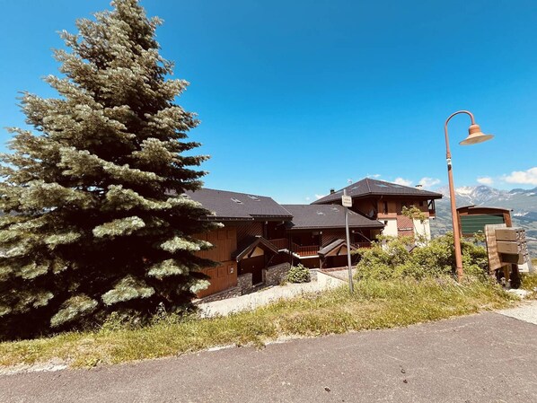 A traditional wooden chalet with a steep roof, surrounded by greenery and a clear blue sky.