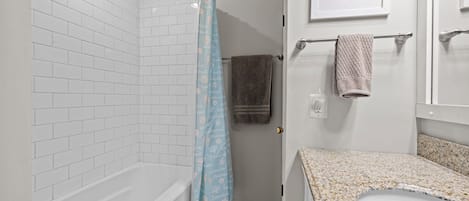 A bathroom with a white tiled shower, a bathtub, a sink with a granite countertop, and a towel rack with towels.