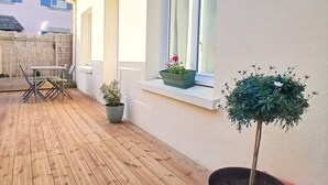 Una terrassa de fusta amb taula i cadires, plantes en testos i una finestra que mostra edificis veïnals.