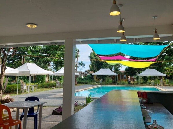 Un espace piscine extérieur doté d’un bar, de parasols blancs et de voiles d’ombrage en tissu aux couleurs vives.