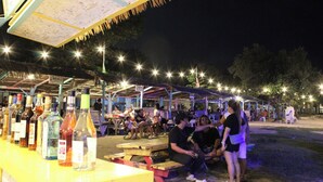 An outdoor bar area with tables and chairs, bottles of beverages, and people sitting and standing. - The Beach Park hadsan by Hiverooms (Lapu-Lapu)