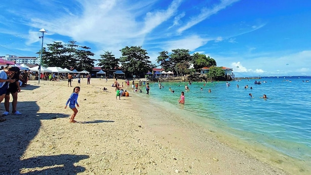 A beach with clear blue water, people swimming and walking on the sand, and some tents in the background. - The Beach Park hadsan by Hiverooms (Lapu-Lapu)