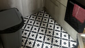 Cocina compacta con encimera negra, pileta de acero inoxidable y microondas. Hay alacenas con cajones y un bote de basura. El piso tiene un estampado geométrico en blanco y negro.