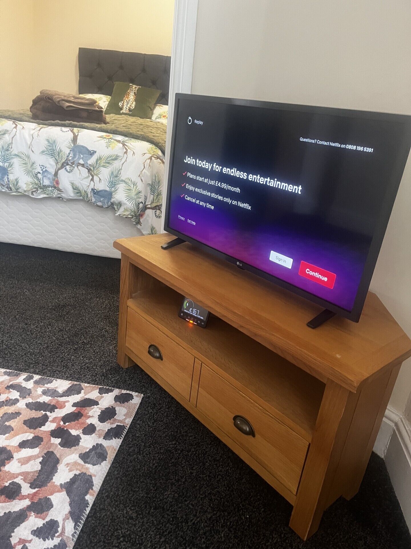A television on a wooden stand with a remote control drawer below it, placed in a room with a bed and a patterned rug.