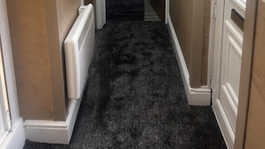 Un pasillo estrecho con alfombra negra que conduce a una zona de cocina con refrigerador, microondas y armarios.