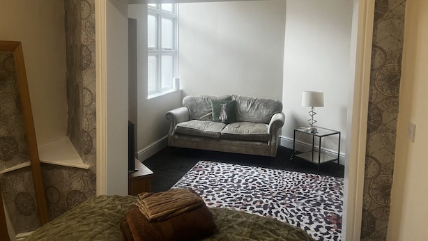 Una habitación con papel tapiz estampado, un sofá gris, un alfombra de estampado de leopardo y una pequeña mesa con una lámpara.