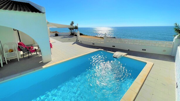Una piscina con vistas al mar, un área de estar cubierta con sillas y un edificio blanco.