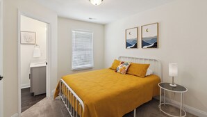 Un dormitorio con cama, mesita de noche, lámpara, ventana con persianas, puerta que da a un baño y dos cuadros colgados en la pared.
