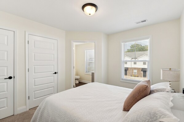 A bedroom with a large bed, white walls, a window with blinds, and a ceiling light.