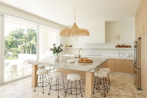 A modern kitchen with a large island, bar stools, a hanging light fixture, and a view of greenery outside.