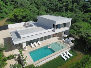 A modern two-story house with a rooftop terrace, a swimming pool, and a lush green forest backdrop.