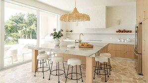 Cocina moderna con una isla grande, taburetes de bar, una lámpara colgante y vistas al exterior con vegetación.