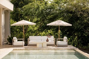 A poolside seating area with two white sofas and two matching umbrellas, surrounded by lush greenery.