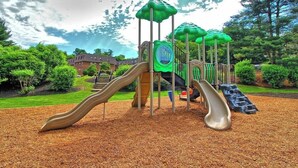 Un parque infantil con toboganes y estructuras para escalar, rodeado de un jardín ajardinado y un edificio al fondo.