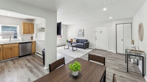 Una cocina moderna con armarios de madera y lavavajillas, un comedor con mesa y sillas de madera, y una sala de estar con un sofá gris y una mesa de centro.