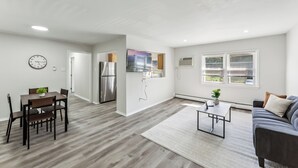 Una sala de estar moderna con un sofá gris, un comedor con una mesa y sillas de madera, una cocina con electrodomésticos de acero inoxidable y un ventanal con persianas.
