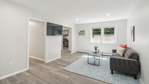 A modern living room with a grey sofa, a coffee table, and a blue rug. There is a TV mounted on the wall and a window with a view of greenery outside.