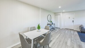 Una habitación moderna y bien iluminada, con una mesa blanca y sillas grises, una mesita auxiliar con una planta, y un sofá gris.