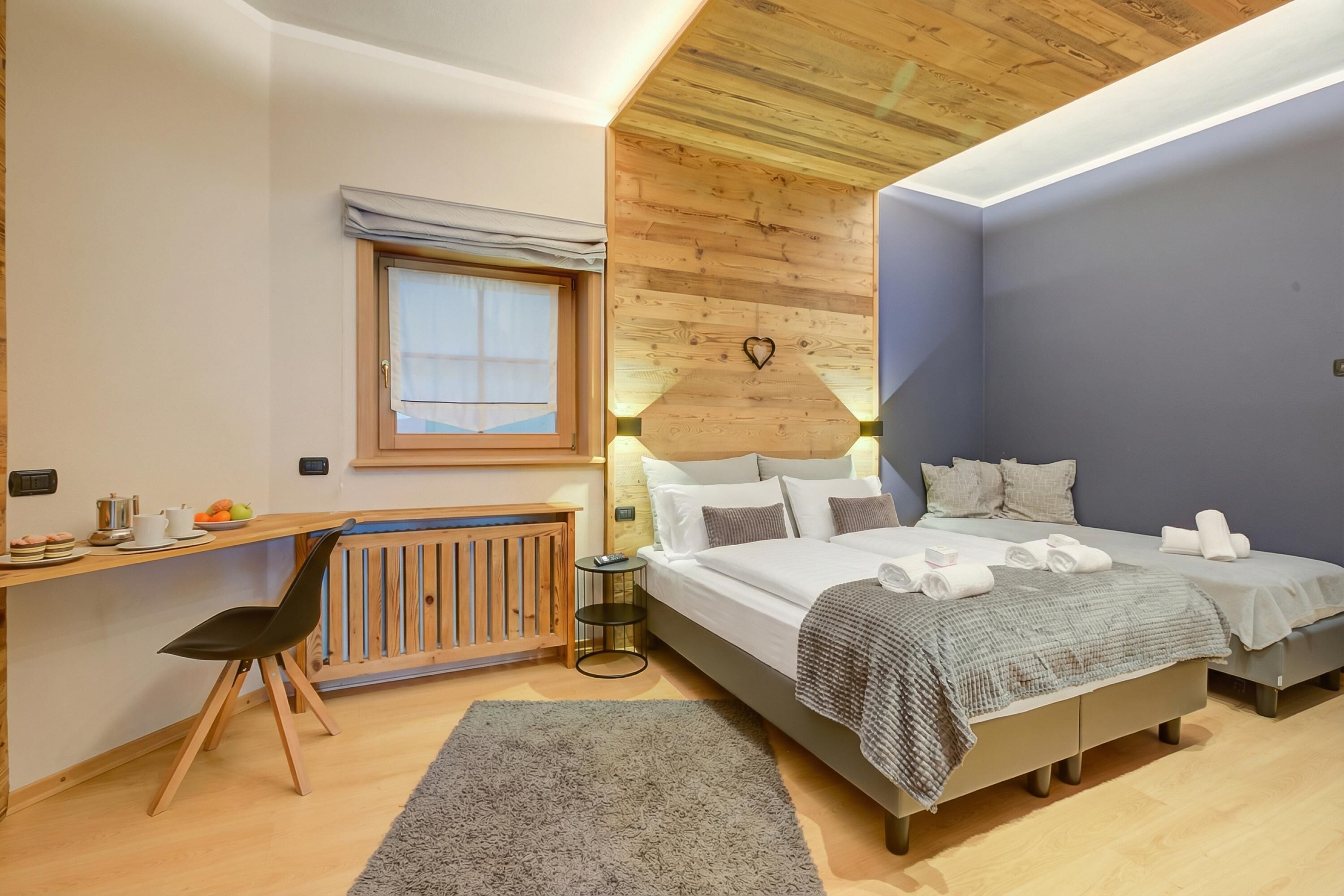 A bedroom with a wooden ceiling, a desk with a chair, a bed with two pillows, and a small bedside table.