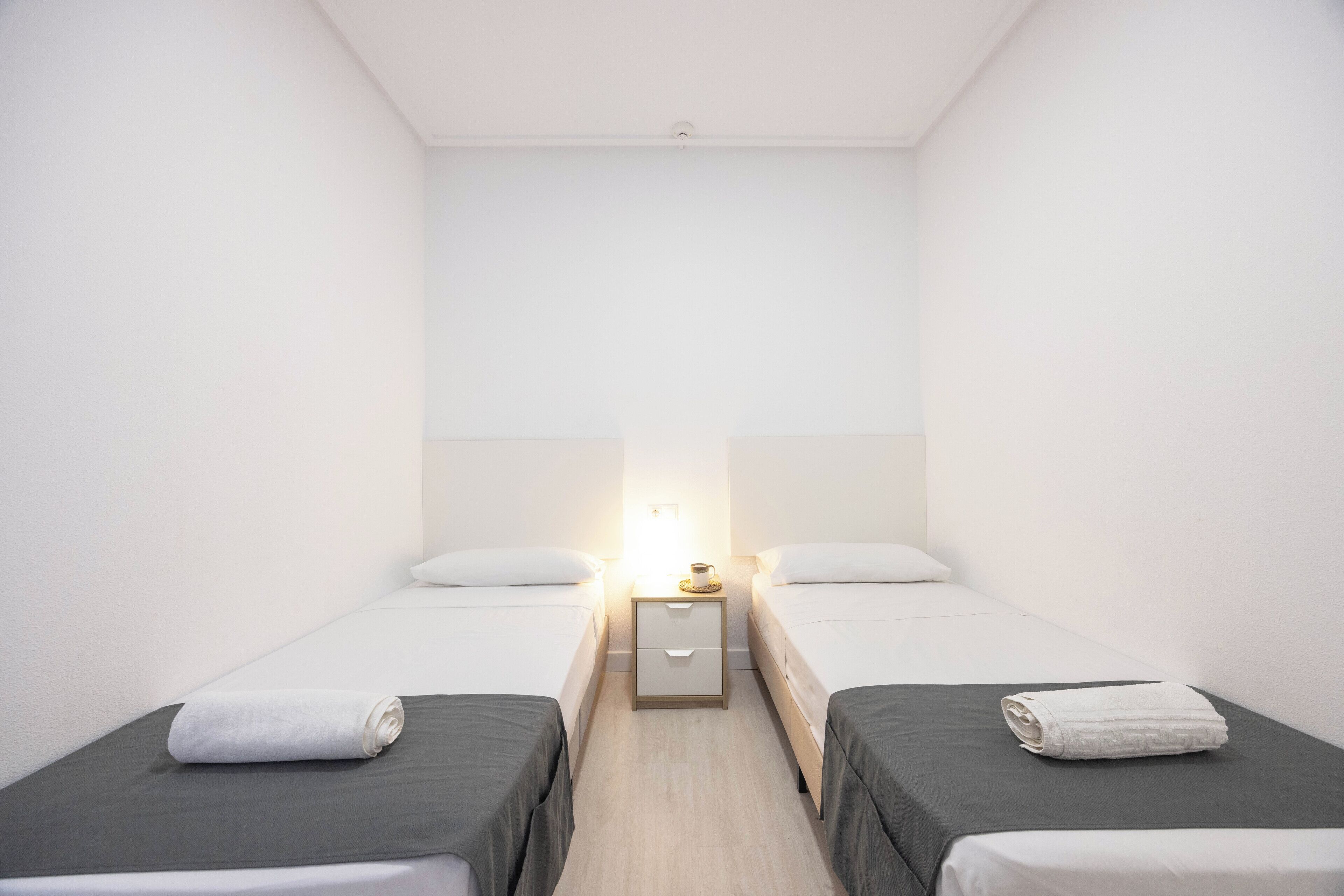 Habitación doble con dos camas individuales, una mesita de noche con lámpara y una pequeña mesita auxiliar.