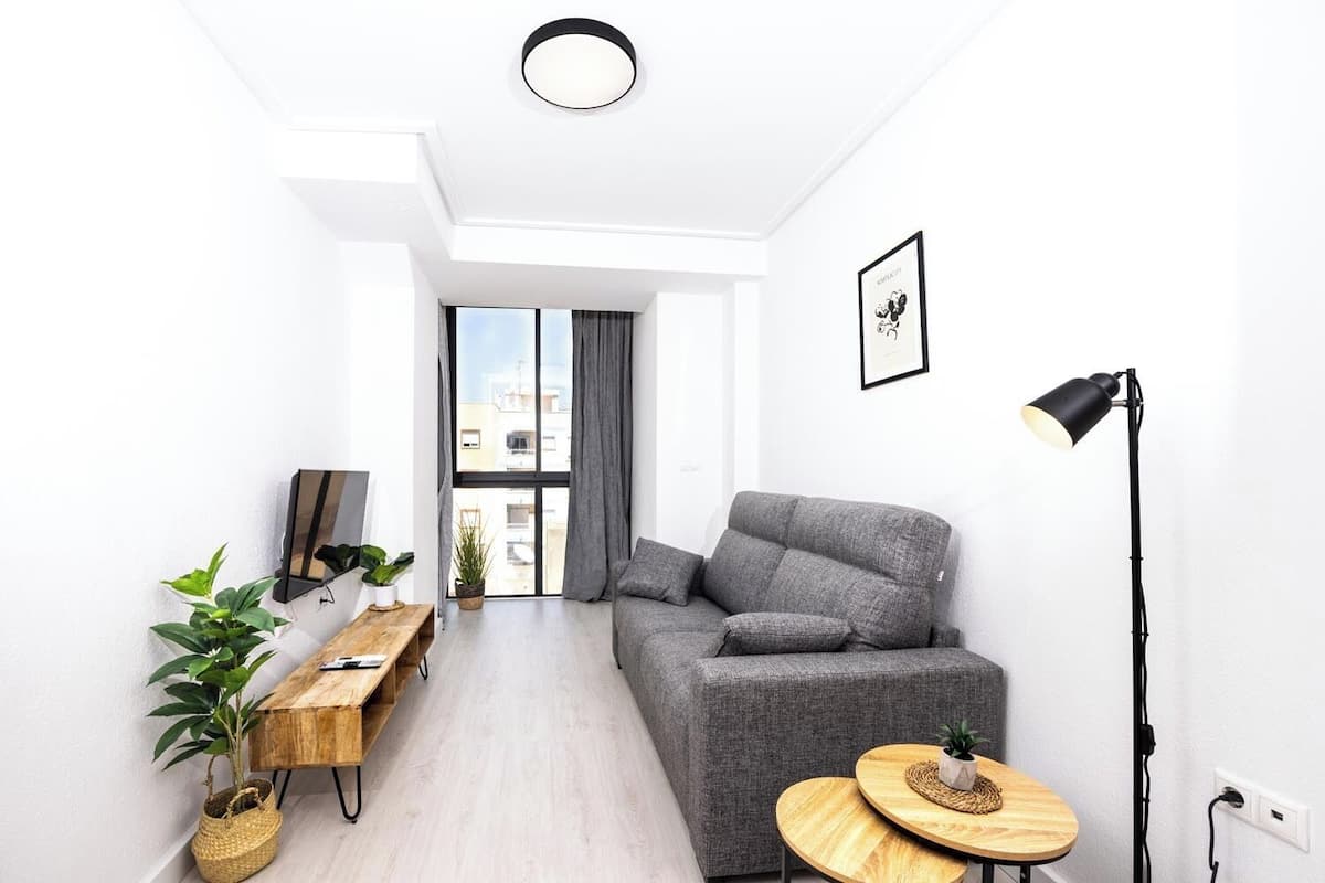 Ein modernes Wohnzimmer mit einem grauen Sofa, einem Holztisch, einer Stehlampe und einem großen Fenster mit Vorhängen.