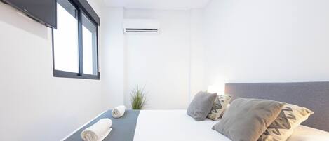 A modern hotel room with a neatly made bed, a window with blinds, an air conditioning unit, and a small plant.