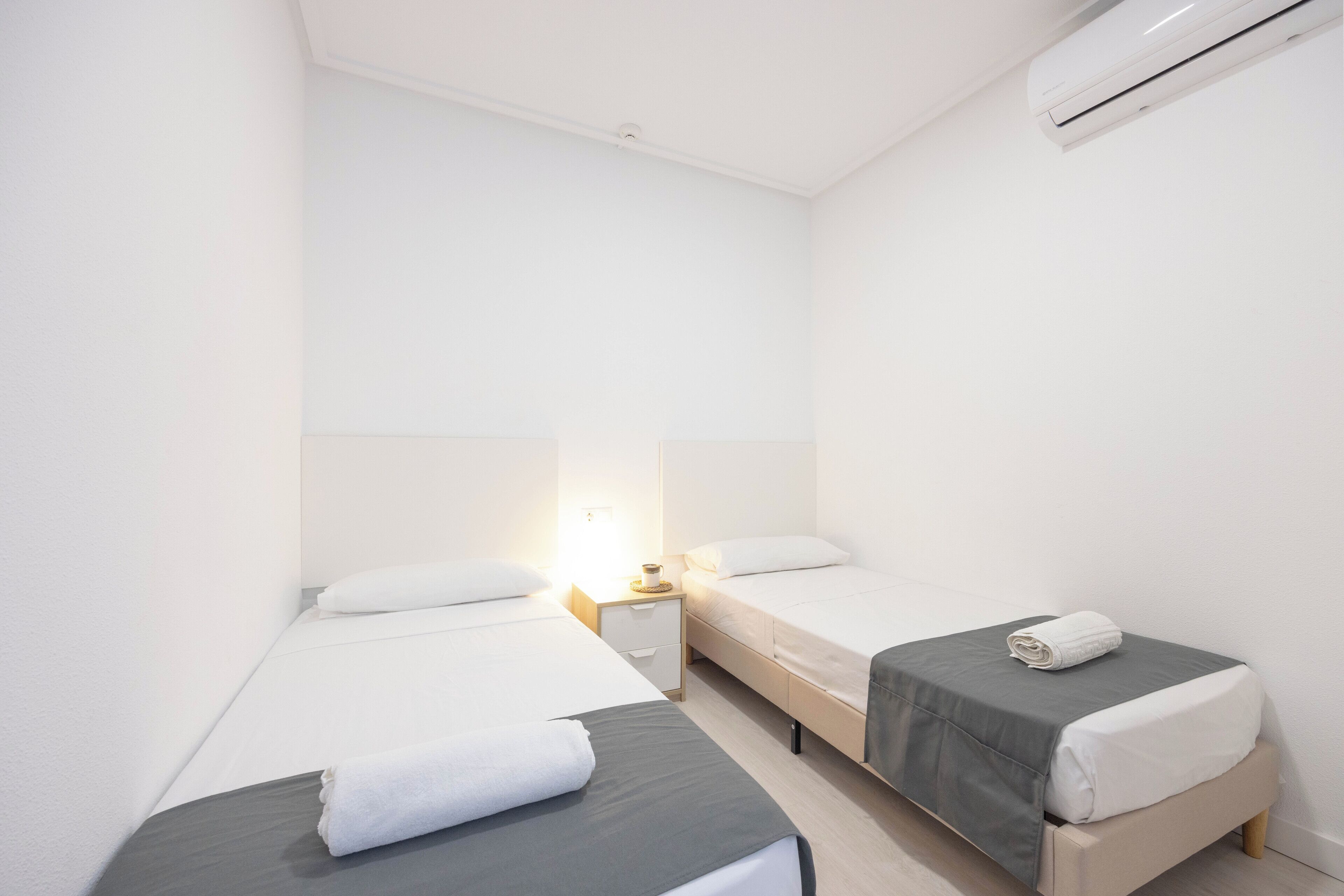Une petite chambre propre, équipée de deux lits simples, d’une table de chevet et d’un climatiseur fixé au mur.