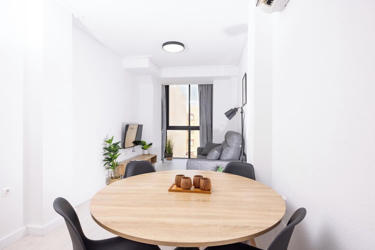 Una habitación moderna y bien iluminada con una mesa redonda de madera, sillas negras, un sofá gris y una ventana con cortinas.