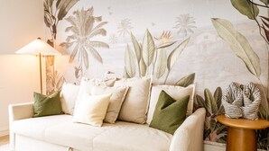 Een woonkamer met een beige bank, groene en witte kussens, een rond houten bijzettafeltje en een muurschildering van tropische planten.