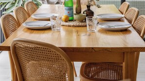 A dining area with a wooden table set for four, surrounded by wicker chairs, with a view of greenery through large windows.