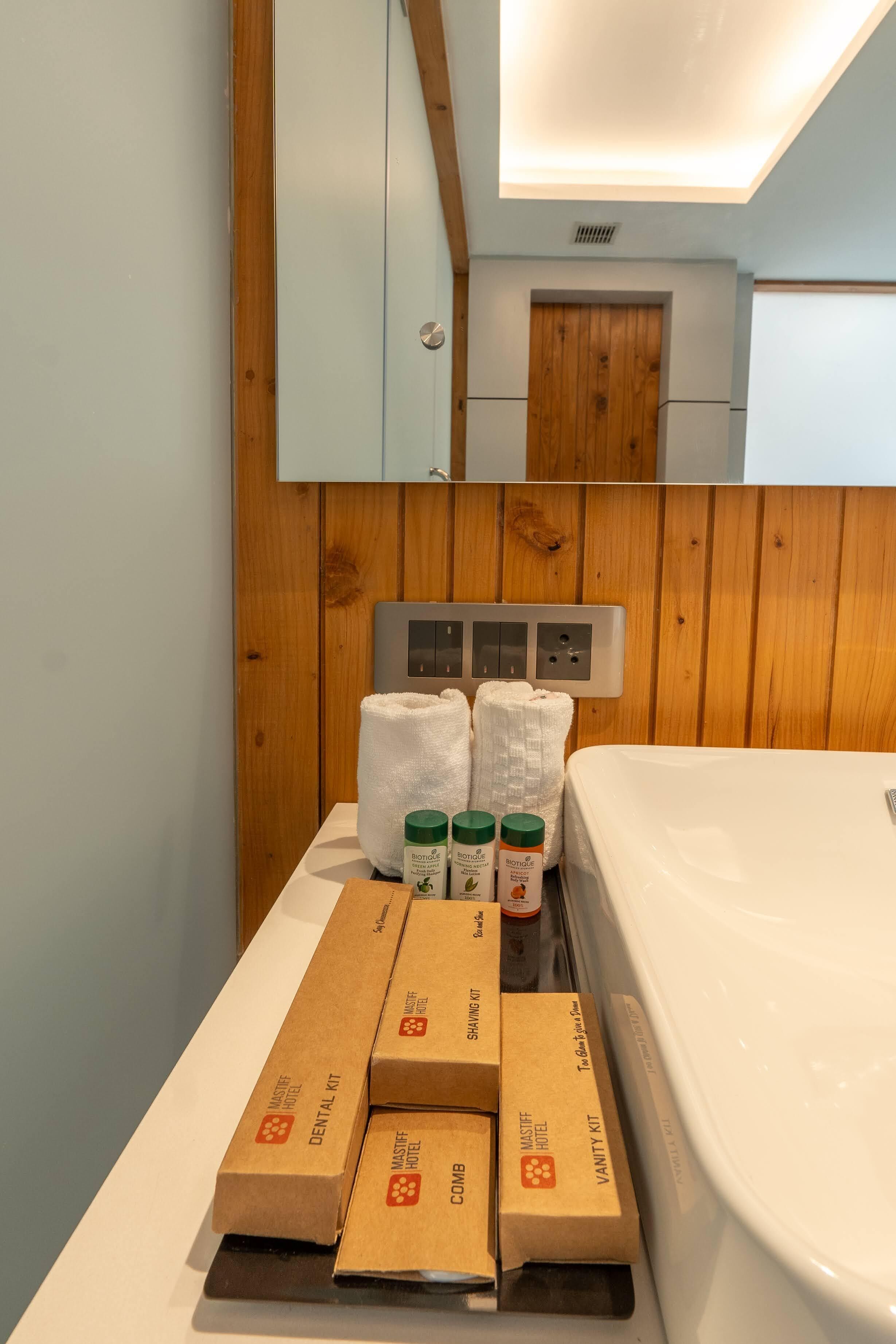 Quarto de hotel com parede de painéis de madeira, cama branca e mesa com artigos de higiene pessoal e kit de barbear.