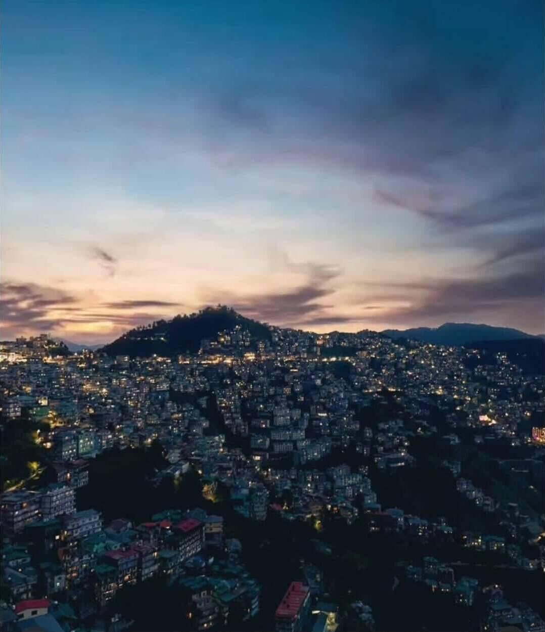 A cityscape at dusk with a densely packed urban area and a hill in the background.