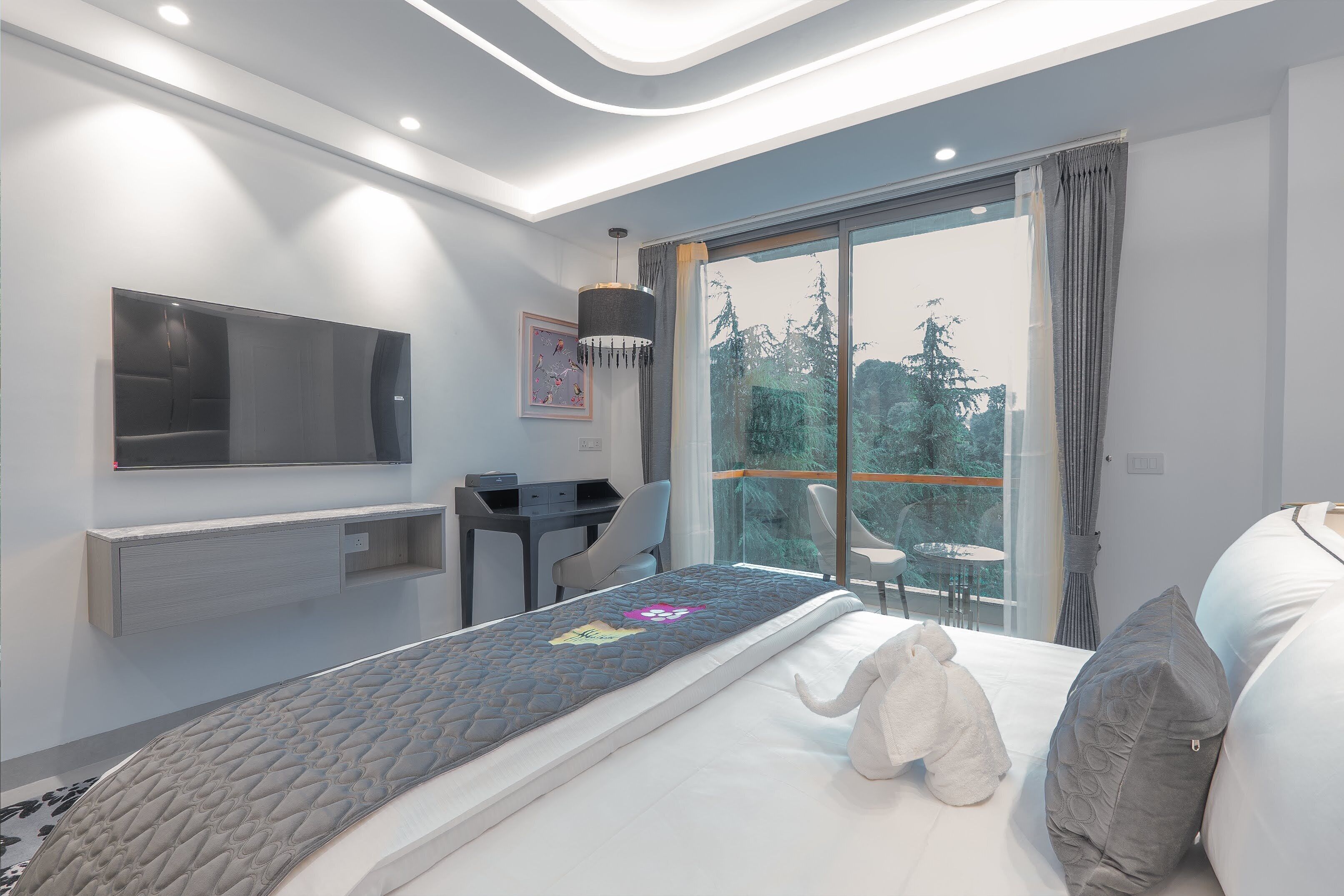 A modern hotel room with a bed, a desk, a chair, and a large window offering a view of trees.