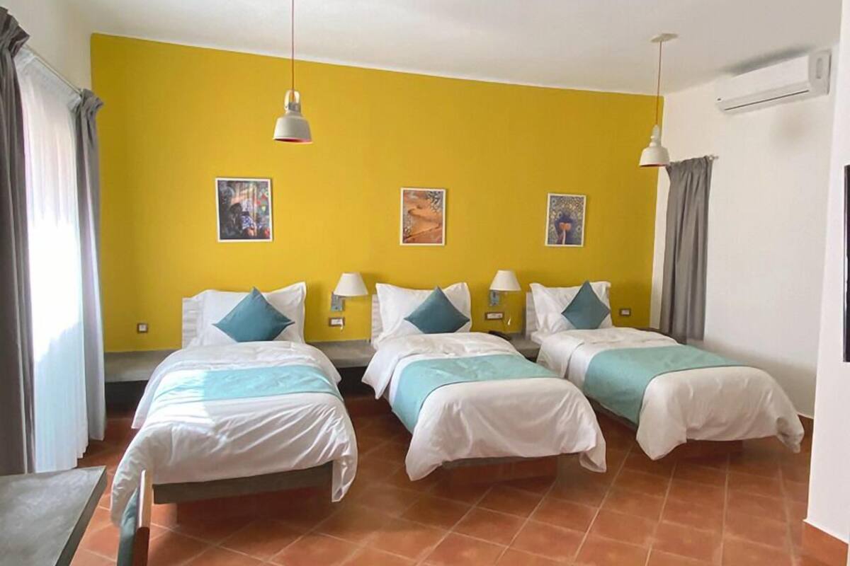A hotel room with three beds, each with white bedding and teal pillows, set against a yellow wall. There are two bedside lamps and three wall-mounted light fixtures. The room has a tiled floor and a window with curtains.