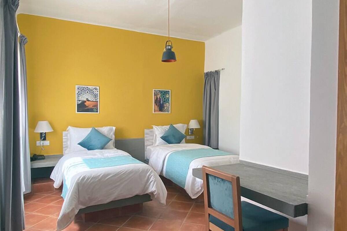 A room with two beds, a desk, and a chair. There are two lamps on the nightstands.