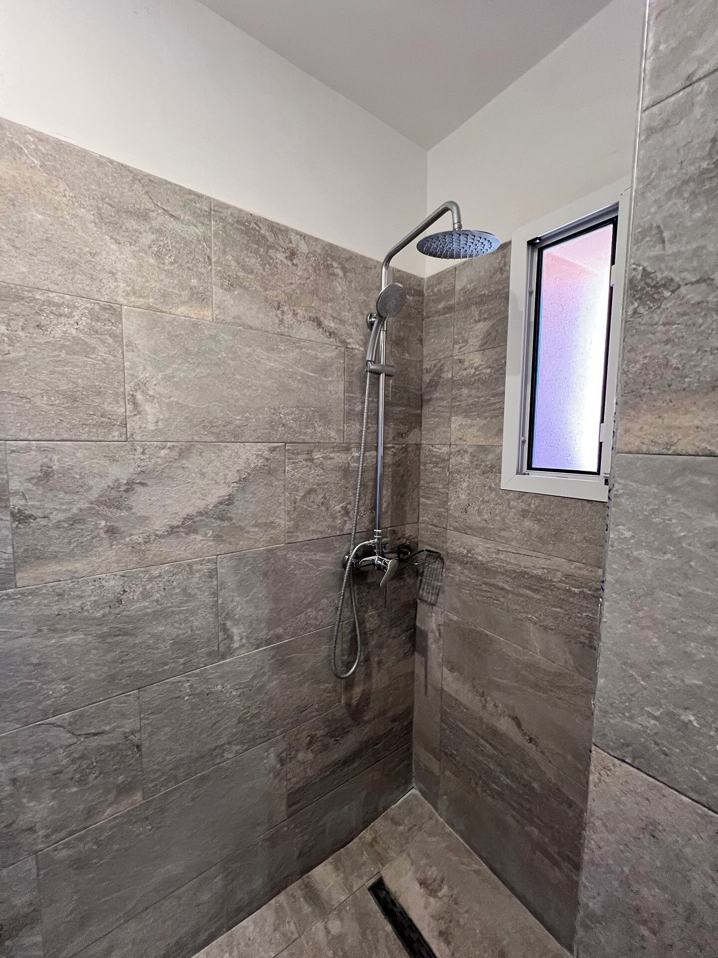 A shower with a handheld showerhead and a wall-mounted faucet, tiled walls, and a small window.